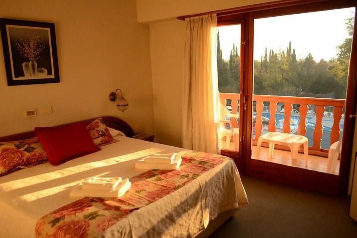 A hotel room with a bed, a painting, a window with curtains, and a balcony with a table and chairs.