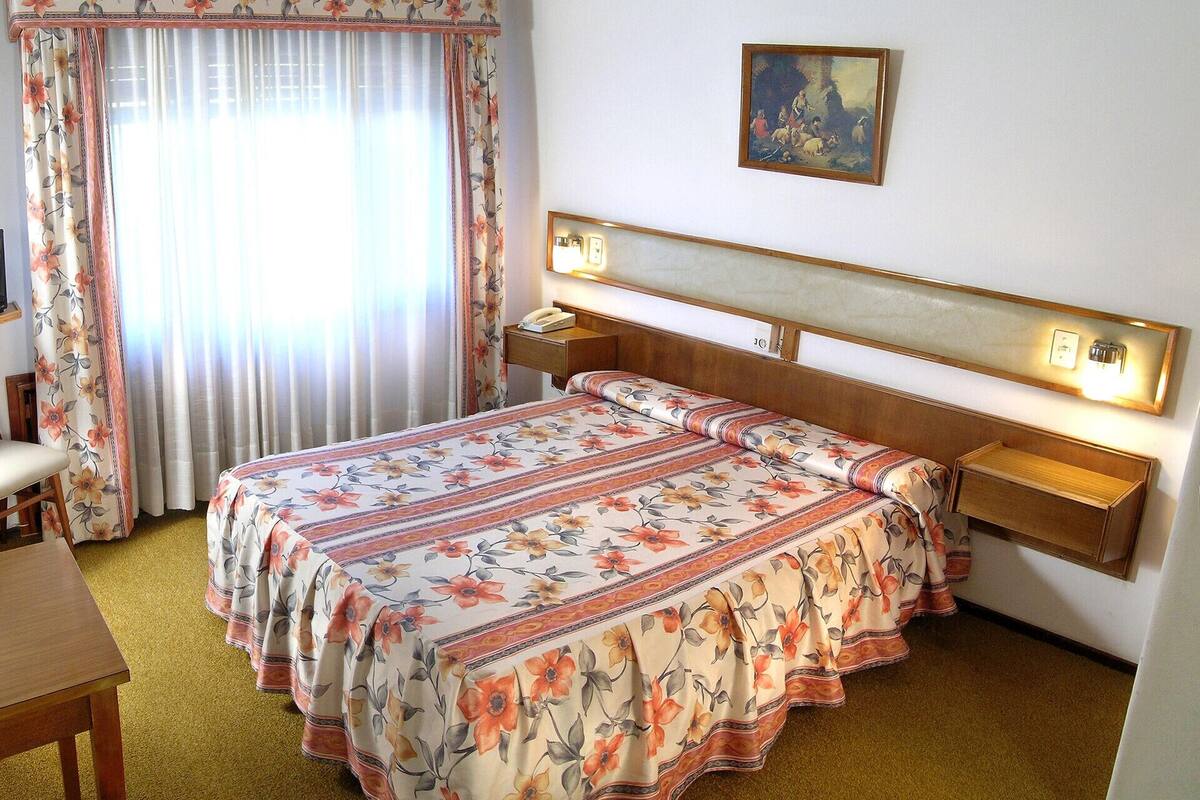 A hotel room with a bed, a desk, a chair, a nightstand, and a painting on the wall.