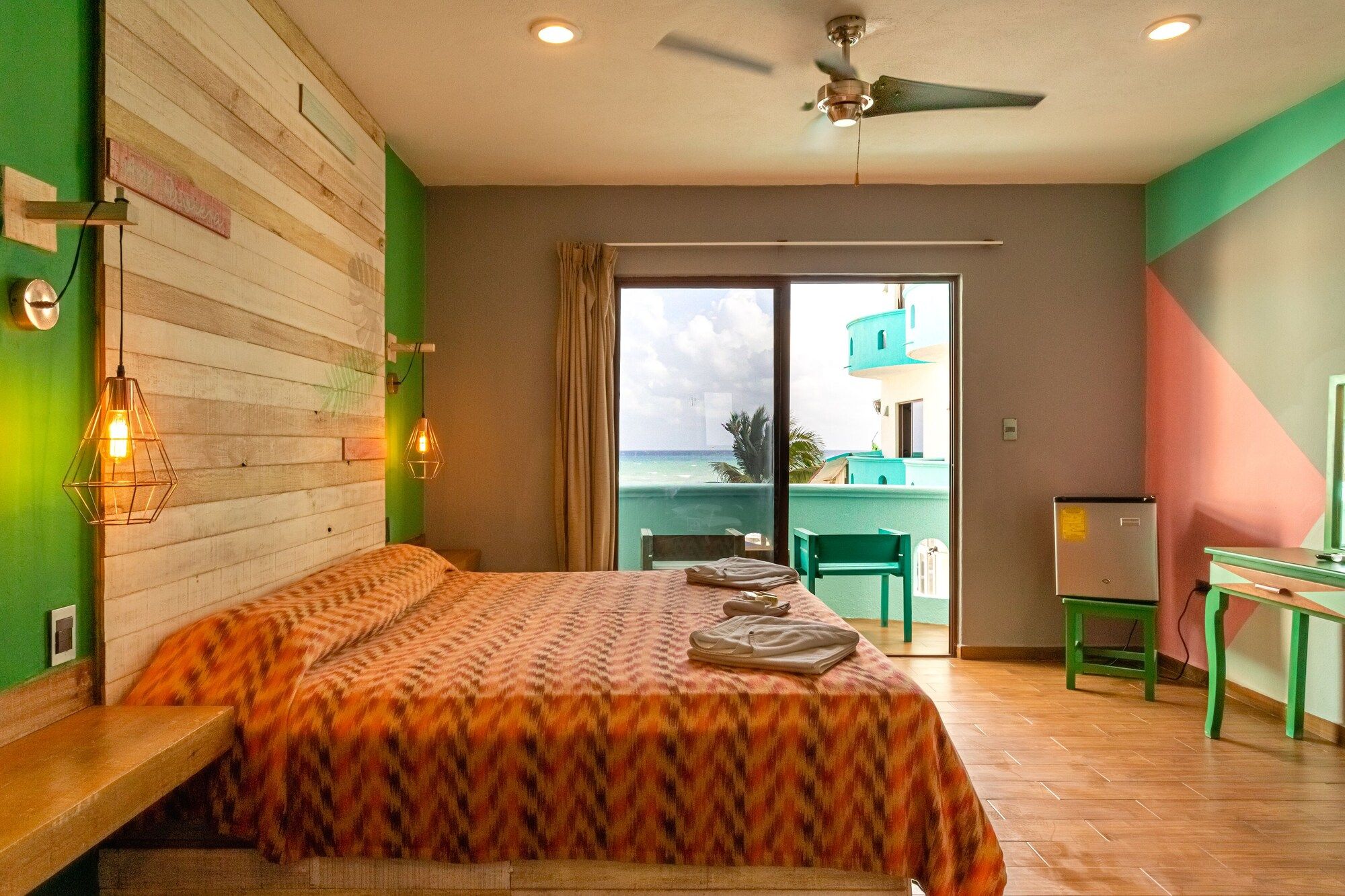 A hotel room with a bed, a ceiling fan, a view of the sea, and a balcony with a table and chairs.