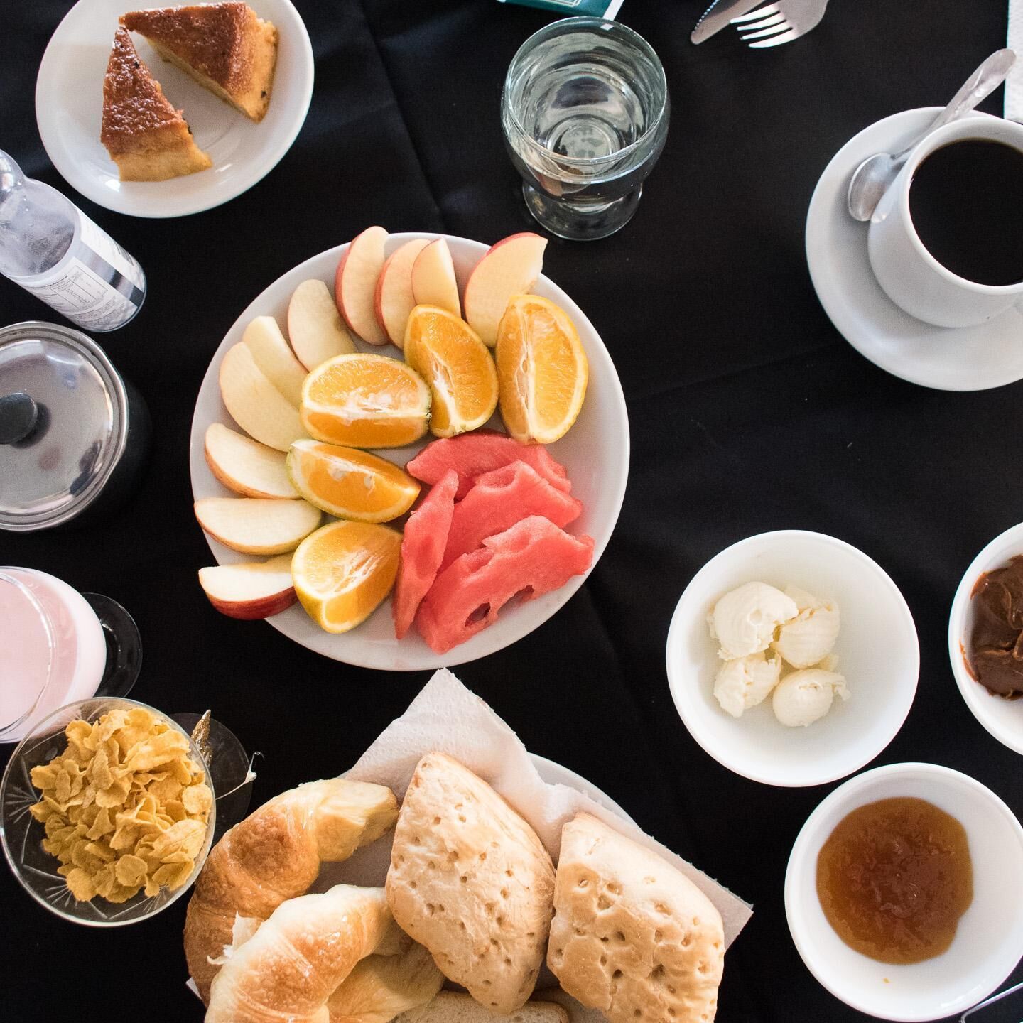 Un desayuno con una fuente de frutas cortadas, un vaso de agua, una taza de café y diversos pasteles.