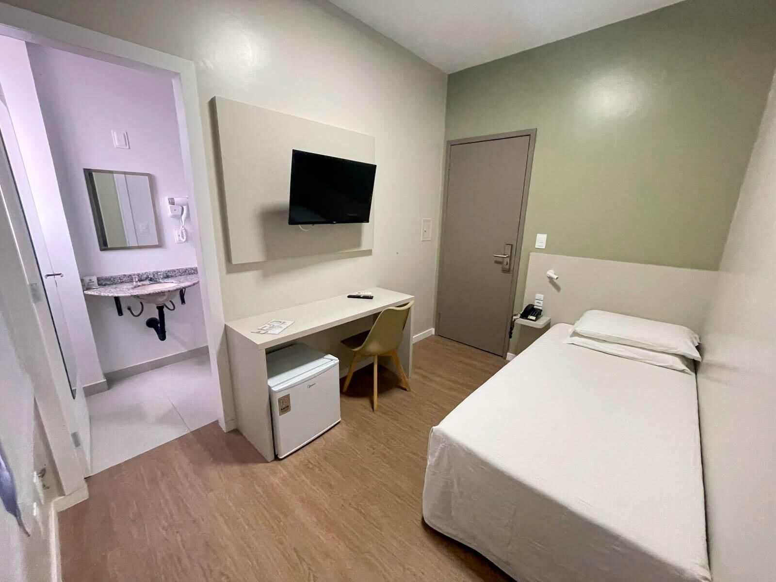 Quarto de hotel com uma cama, uma escrivaninha com cadeira, uma televisão e um pequeno frigorífico.