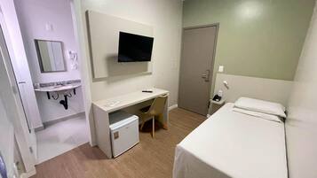 Quarto de hotel com uma cama, uma escrivaninha com cadeira, uma televisão e um pequeno frigorífico.