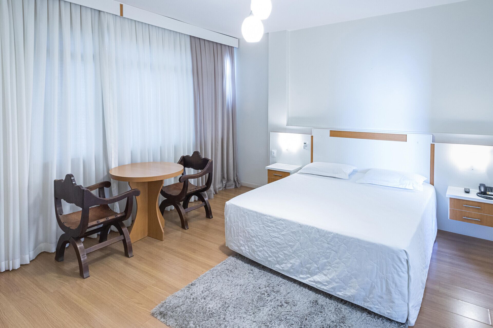 A hotel room with a bed, a round table with two chairs, a small desk with a telephone, and a nightstand with a lamp.