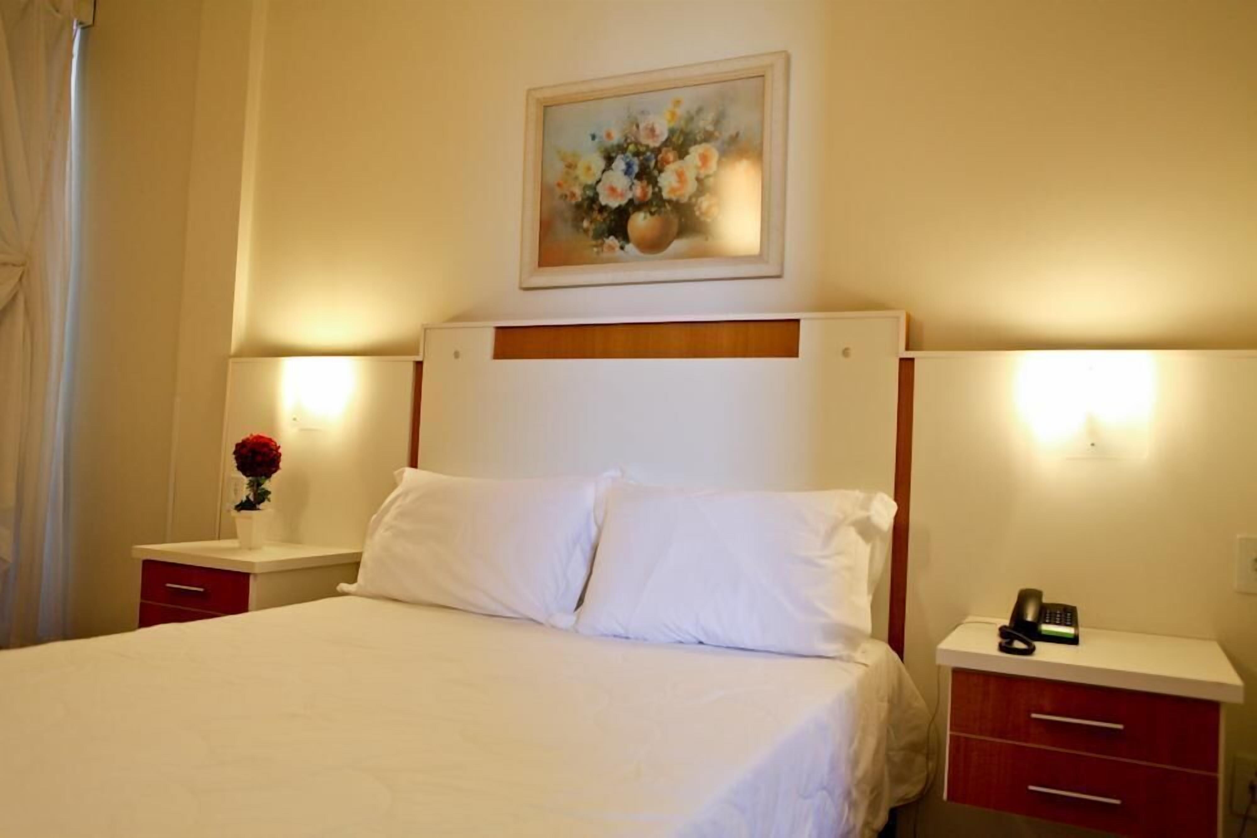Quarto de hotel com uma cama grande, duas mesas de cabeceira com luminárias, um quadro na parede e um vaso com flores em uma das mesas.
