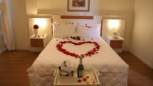 Camera d'albergo con un letto, due comodini, una lampada da comodino, un quadro appeso alla parete e un bouquet di petali di rosa a forma di cuore sul letto.