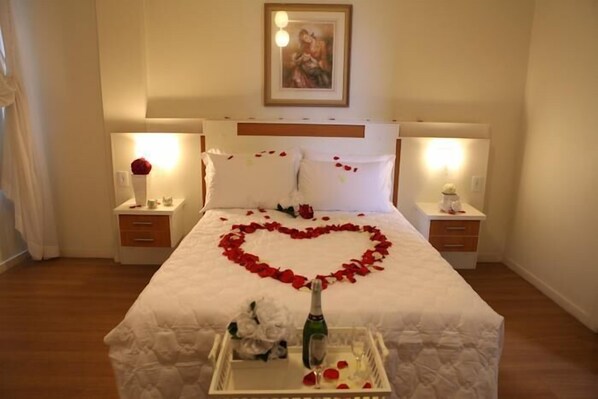 A hotel room with a bed, two bedside tables, a nightstand, a framed picture on the wall, and a heart-shaped arrangement of rose petals on the bed.