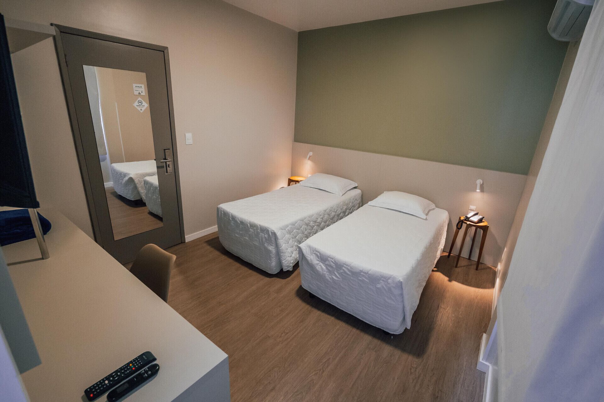 A hotel room with two single beds, a desk with a remote control, a mirror, and a bedside table with a lamp.