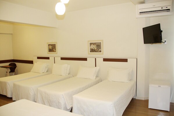A hotel room with three single beds, a small table, two wall-mounted pictures, a TV, and a compact air conditioning unit. - Hotel Gracher (Brusque)