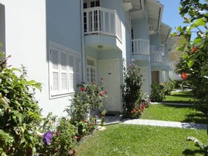A two-story building with white exterior walls, a balcony on the second floor, and a small front garden with various plants and flowers.
