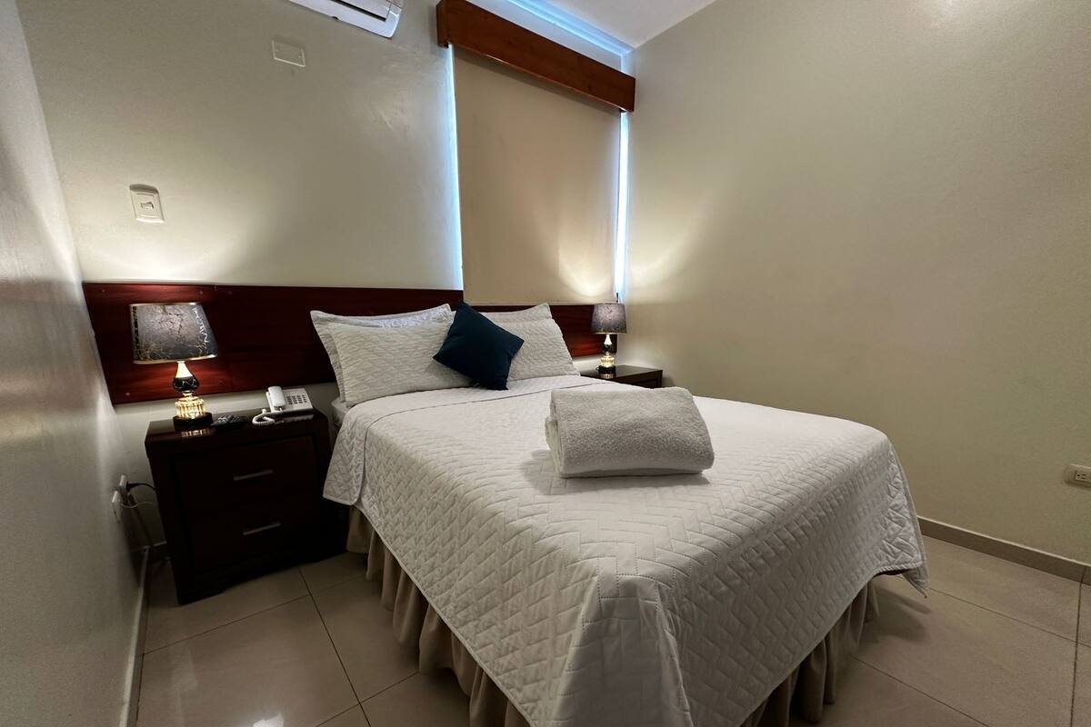 A neatly made bed with a white quilt, a folded towel, and two pillows. There is a nightstand with a lamp and a phone. The room has a wooden headboard and a window with blinds.