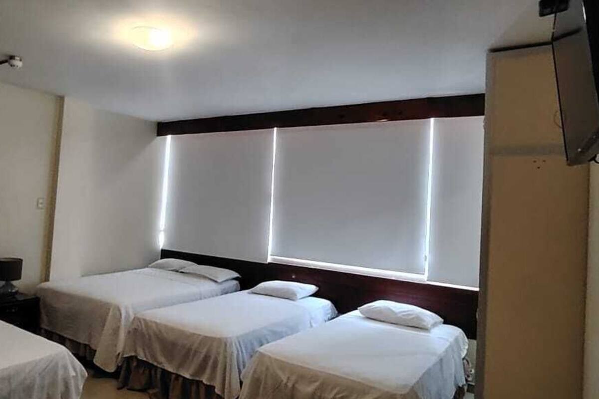 A hotel room with three single beds, a small table with a lamp, and a TV mounted on the wall.