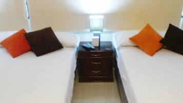 Habitación de hotel con dos camas, cada una con ropa blanca y almohadas anaranjadas y negras. Hay una mesita de noche entre las camas con una lámpara y un teléfono.