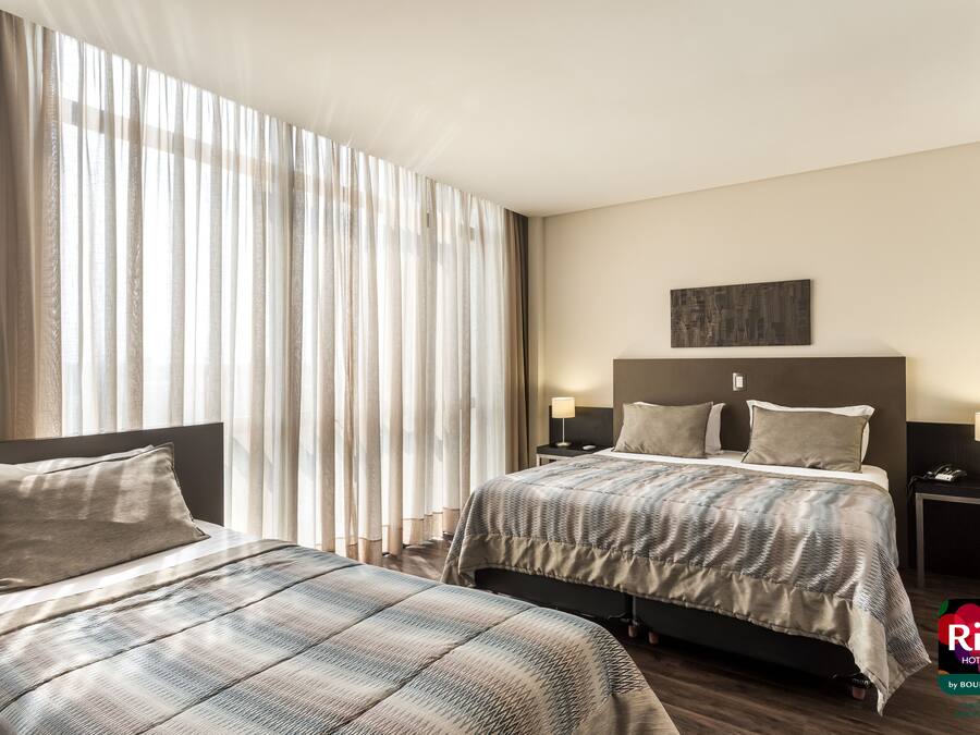 Habitación de hotel con dos camas, un ventanal con cortinas translúcidas, cabecera con un cuadro, lámparas de noche y un mini bar.