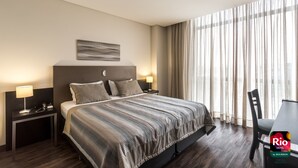 Une chambre d’hôtel avec un grand lit, deux tables de chevet avec des lampes, une chaise et une vue sur l’extérieur à travers des rideaux légers.