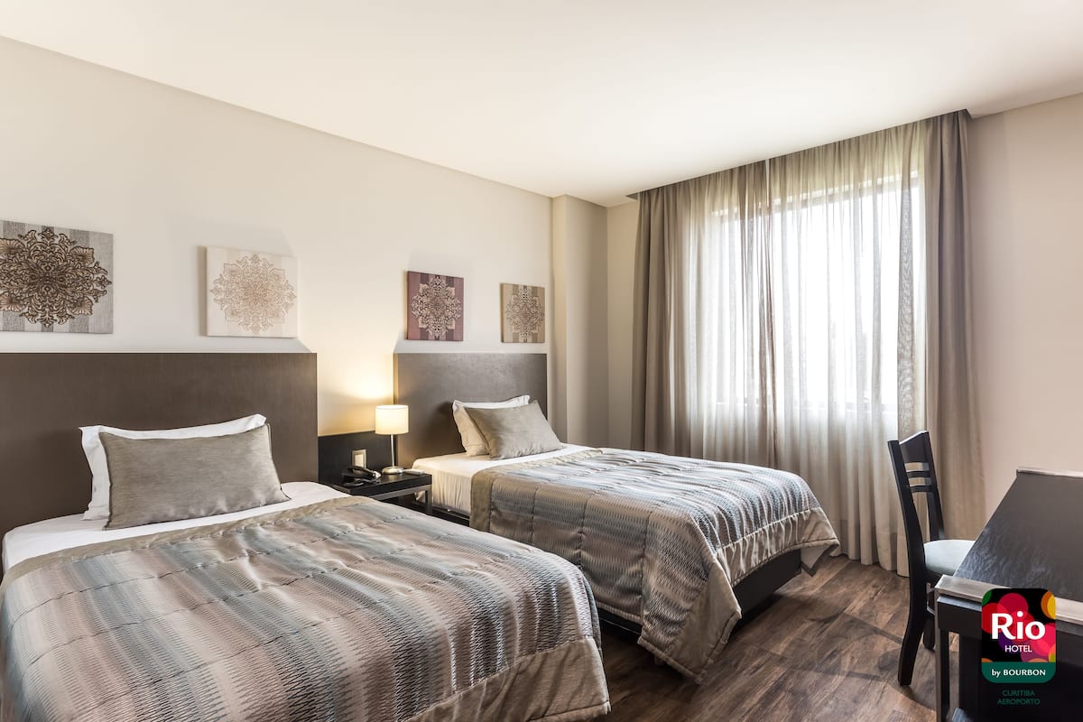 Une chambre d’hôtel avec deux lits, un bureau et une chaise. Des œuvres d’art sont accrochées au mur au-dessus des lits, et il y a une fenêtre avec des rideaux.