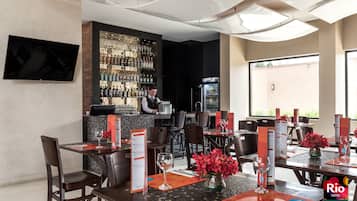 Un restaurante con mesas dispuestas para comer, una zona de bar con un cantinero, y un gran ventanal con vistas.