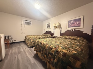 Habitación de hotel con dos camas, un escritorio, una silla, un refrigerador y una ventana con aire acondicionado.