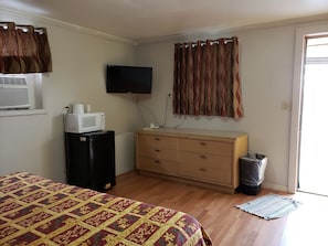 A bedroom with a bed, a dresser, a television, an air conditioning unit, and a waste bin. - Mustang Motel (Gillette)