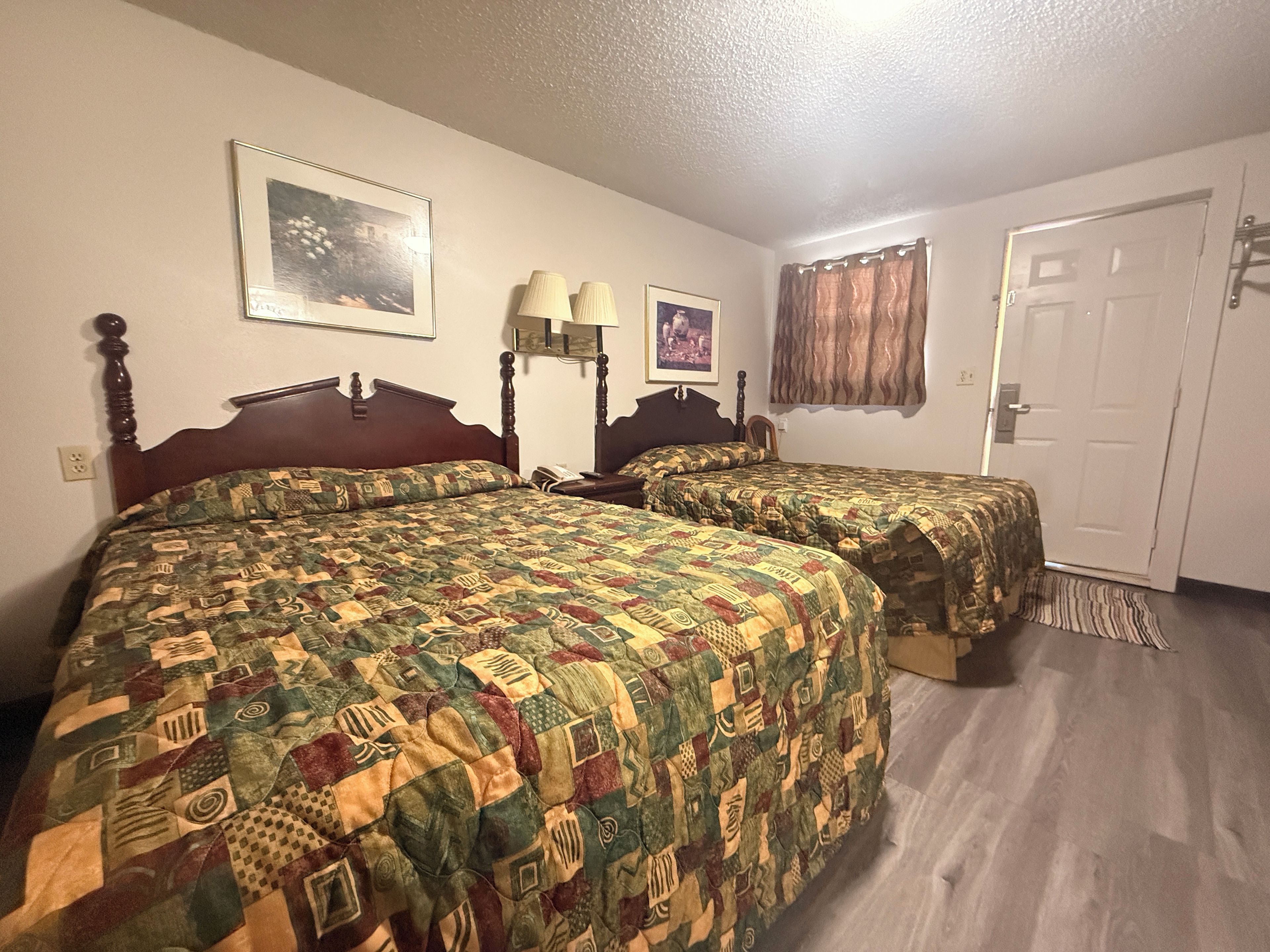 A hotel room with two beds, a door, a window with curtains, and a wall-mounted lamp.