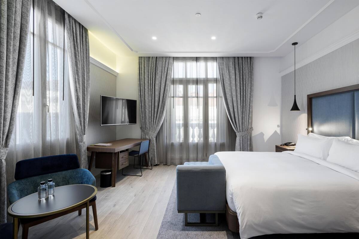 A modern hotel room with a large bed, a desk, a chair, and a TV.