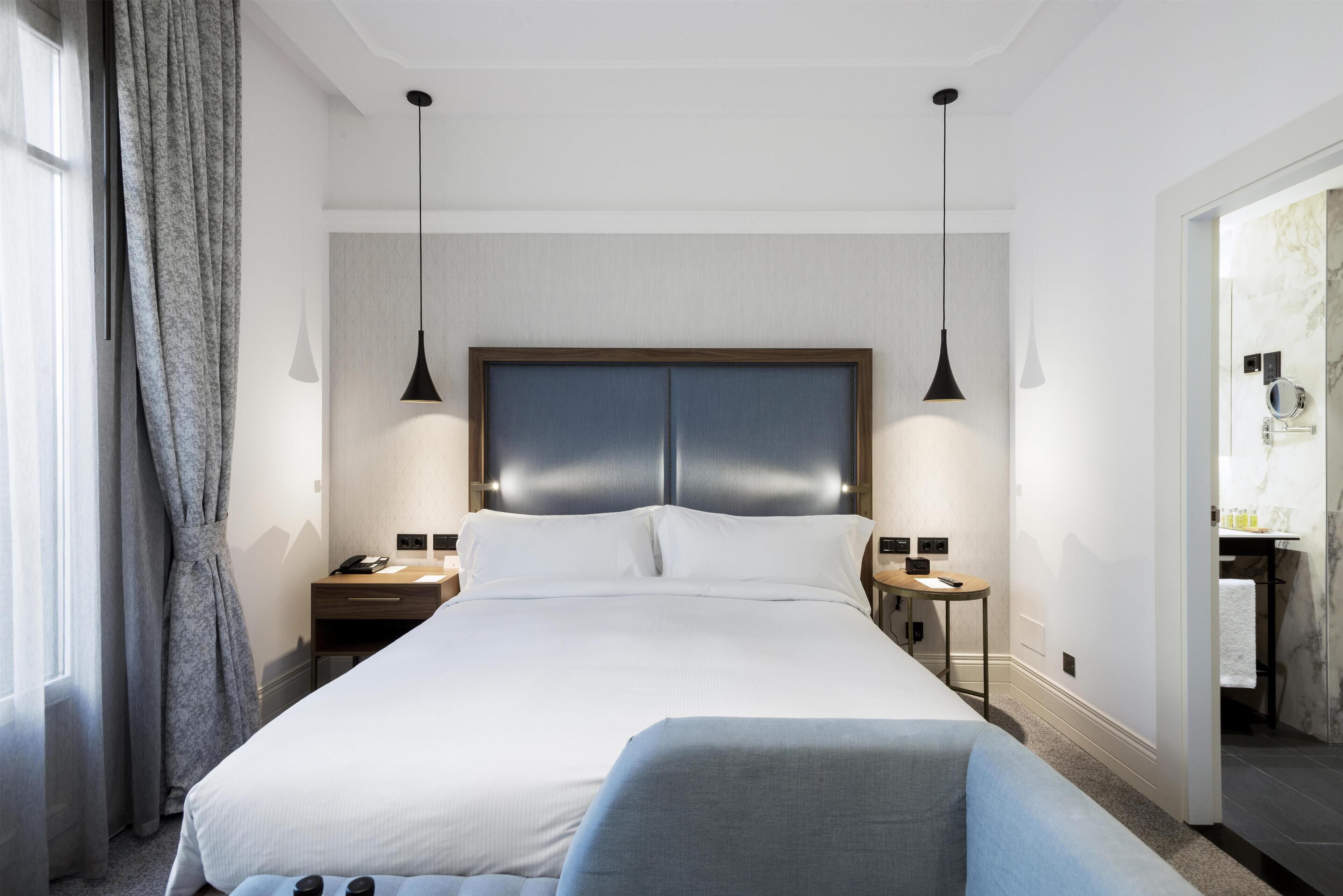 Une chambre d’hôtel moderne avec un grand lit, deux tables de chevet, une petite table, une chaise et une salle de bain visible à travers une porte ouverte.
