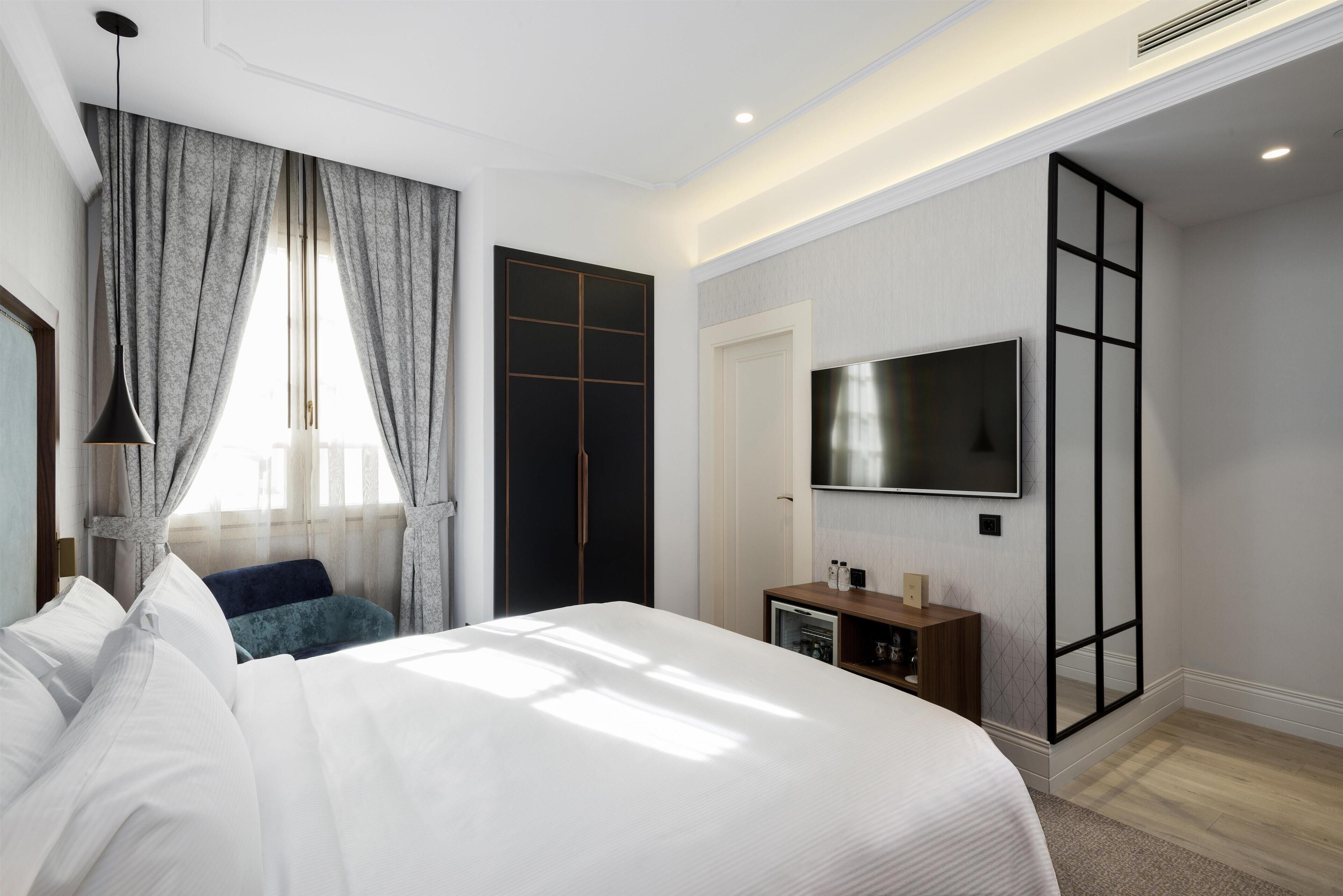 Una habitación de hotel moderna con una cama grande, un televisor montado en la pared, un armario y un escritorio pequeño con una lámpara.