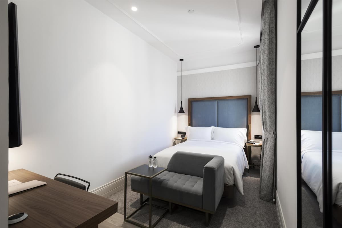 A hotel room with a bed, bedside table, lamp, chair, and a desk with a chair.