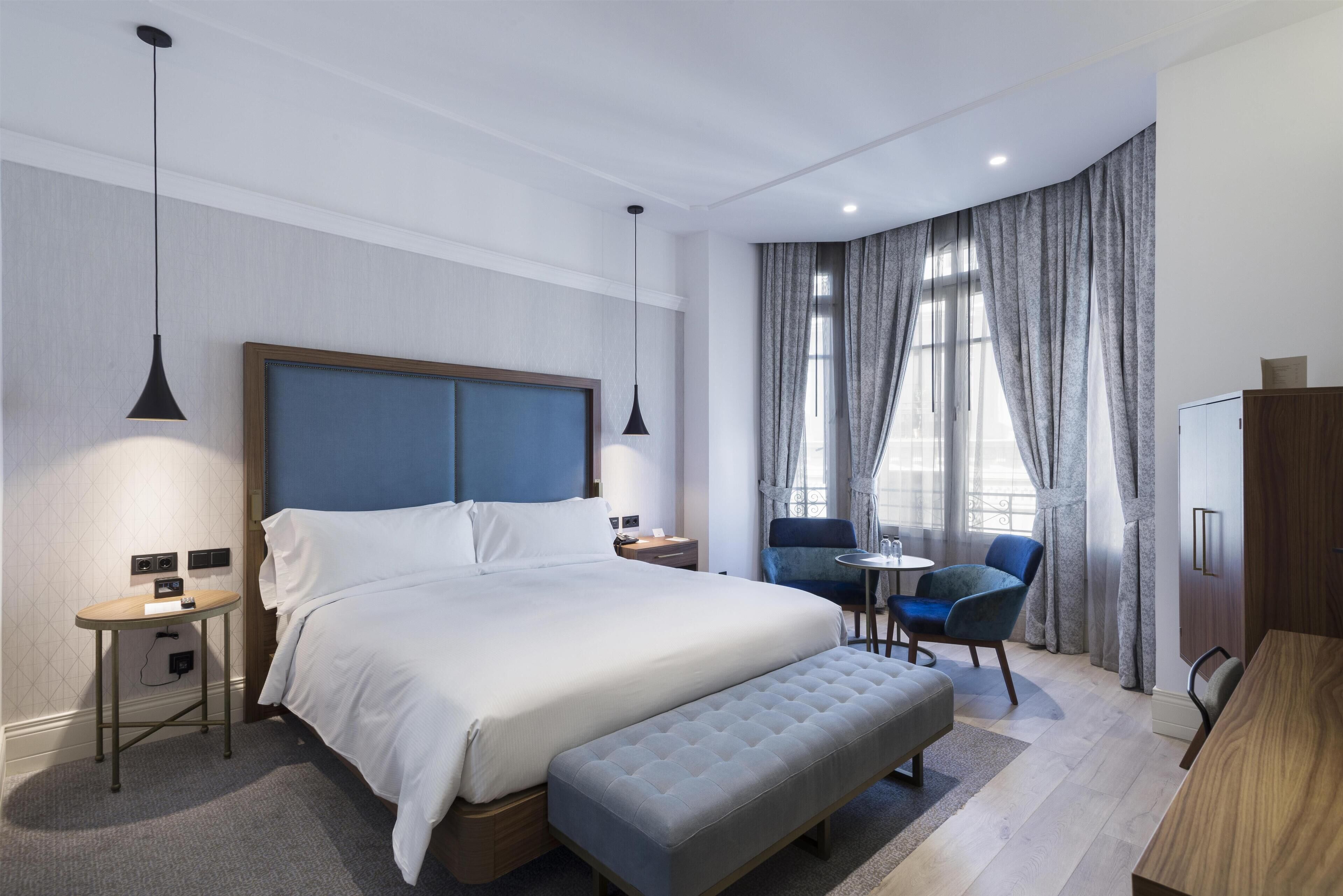 Ein modernes Hotelzimmer mit einem großen Bett, Nachttischen, einem kleinen runden Tisch, einem Sessel, einer Bank und einem Fernseher.
