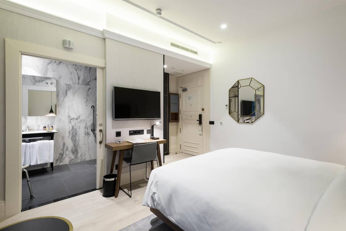 A hotel room with a bed, a desk, a chair, a TV, a mirror, and a bathroom with a shower and sink.
