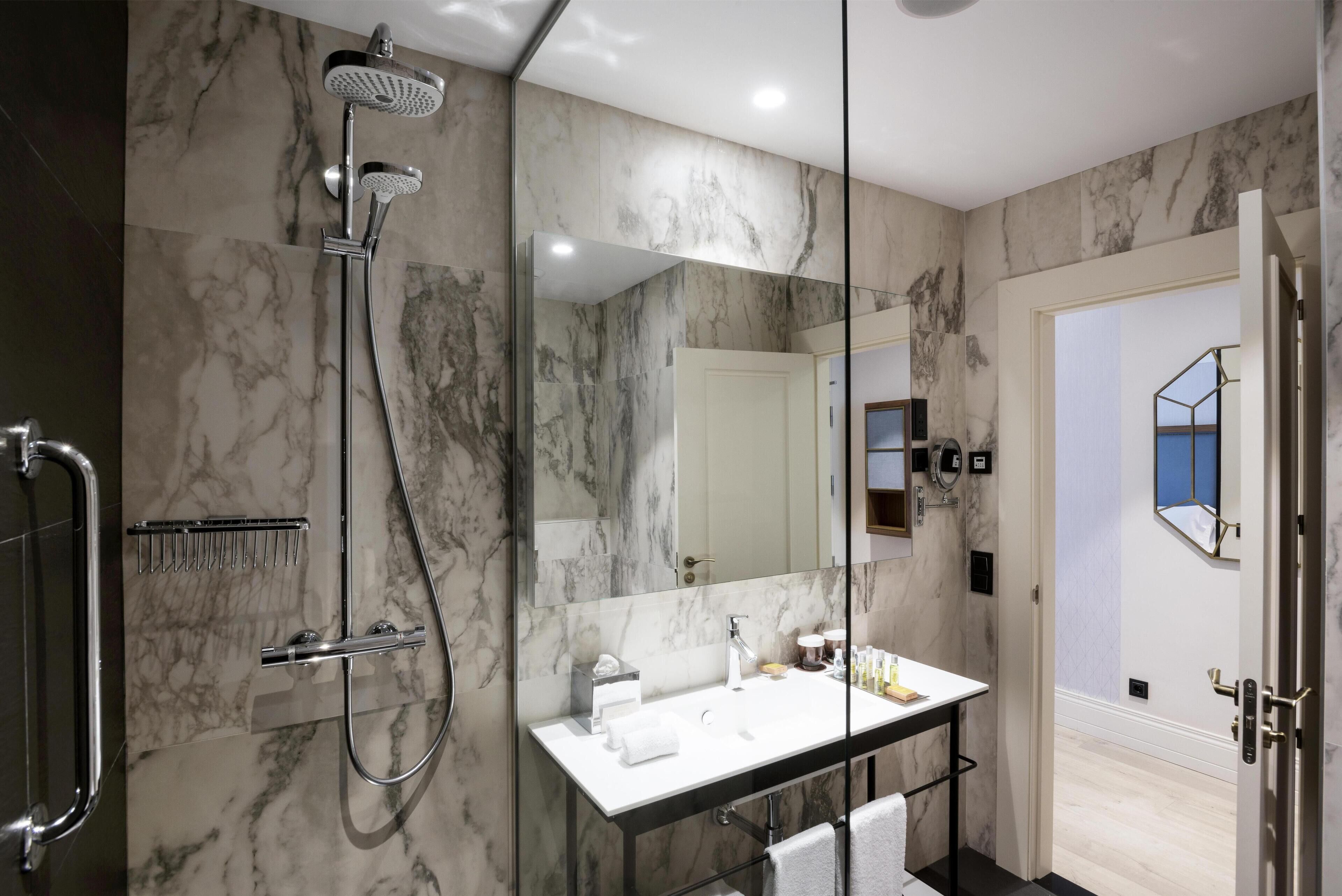 A modern bathroom with a marble wall, a glass-enclosed shower, a white sink, and a large mirror.