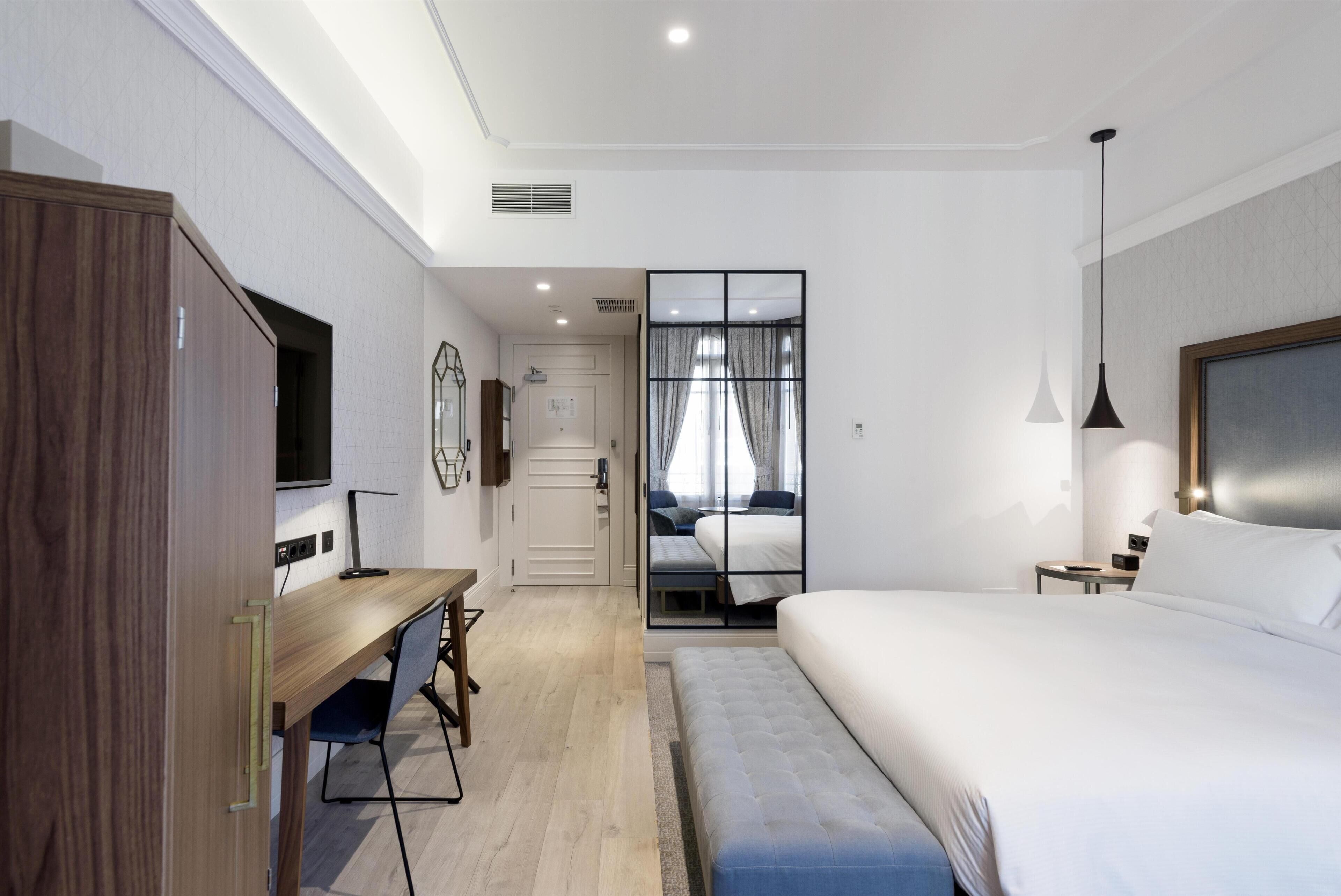 Una habitación de hotel moderna con un escritorio de madera, una cama grande, un televisor y vistas a través de un ventanal.