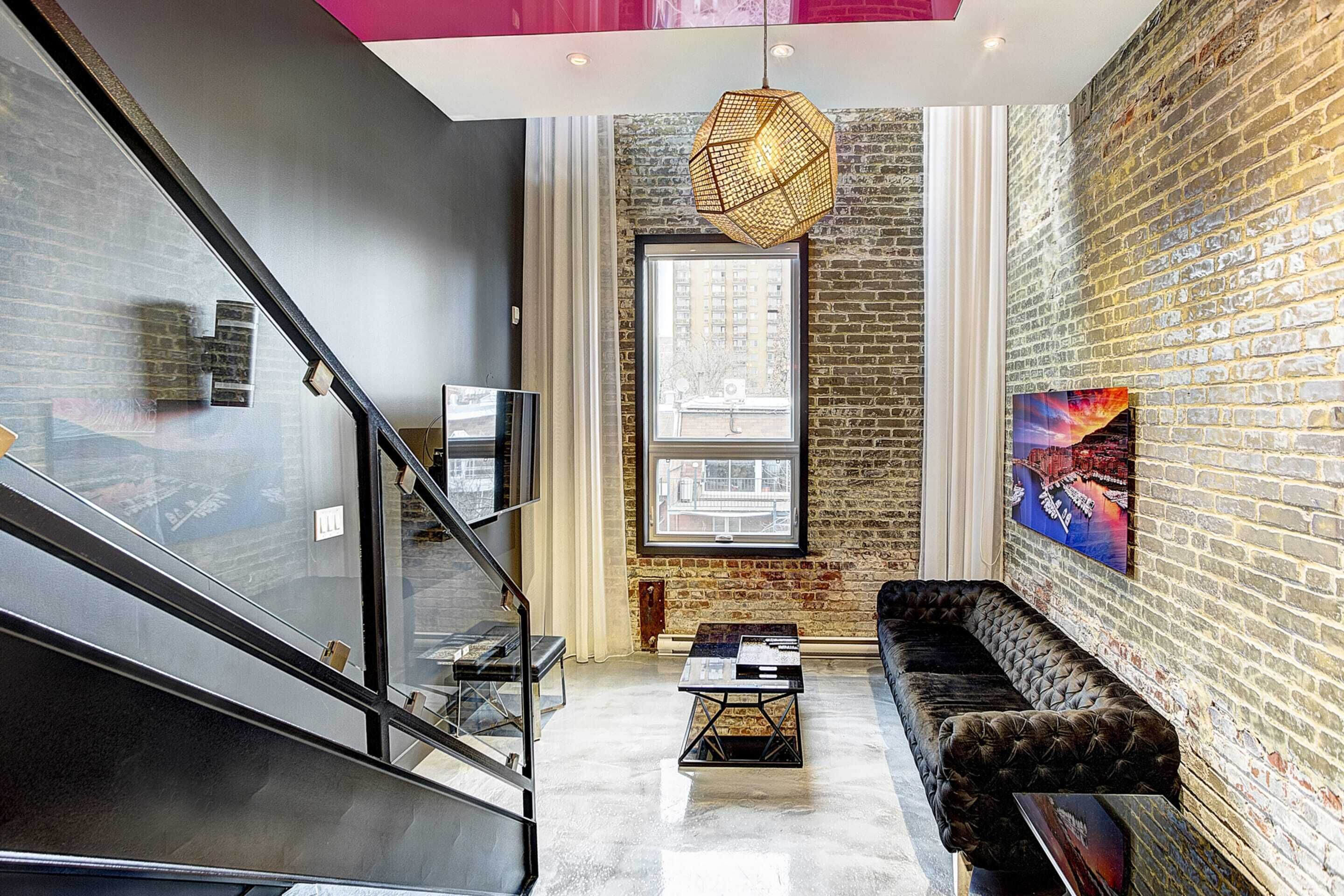 A modern living room with a black staircase, a large window, a black sofa, and a hanging light fixture.