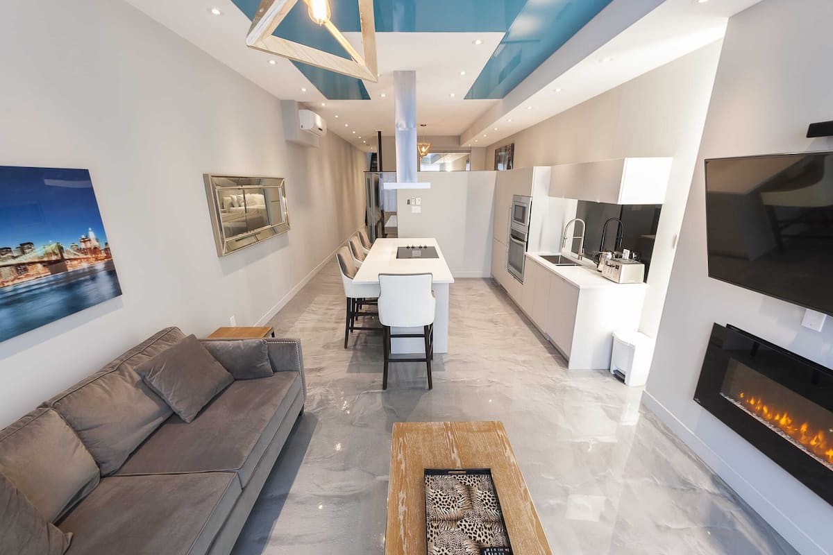 Uma sala de estar moderna com um sofá cinzento, uma mesa de centro e uma área de jantar com mesa e cadeiras brancas. Há também uma cozinha com fogão, pia e uma televisão fixada na parede.
