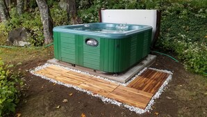 A green hot tub with a control panel, surrounded by a wooden deck and white pebbles, set in a garden with trees and plants. - The Farmstand Bed & Breakfast (Chocorua)