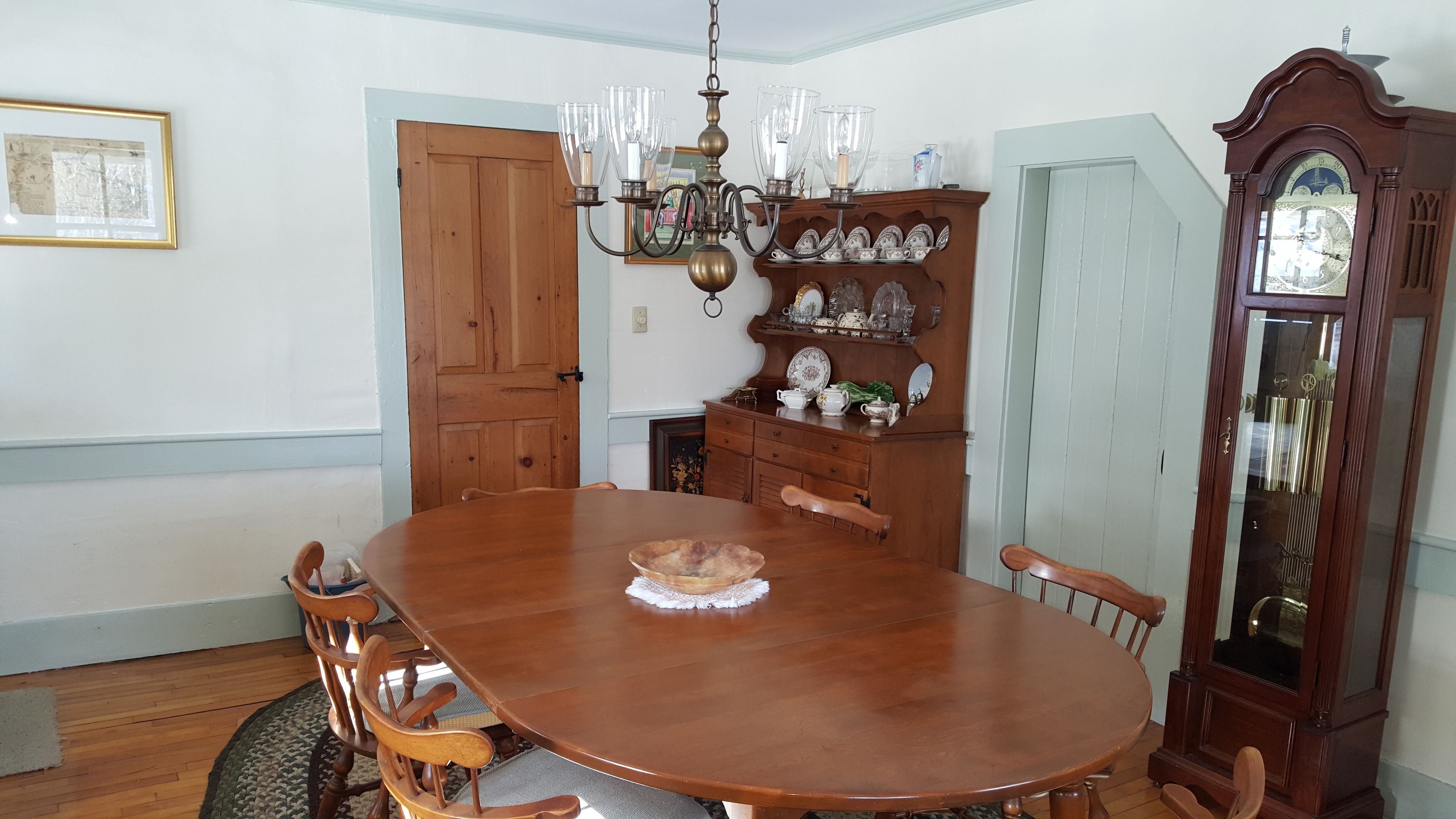 A dining room with a wooden table, chairs, a chandelier, a wooden cabinet with dishes, and a grandfather clock.