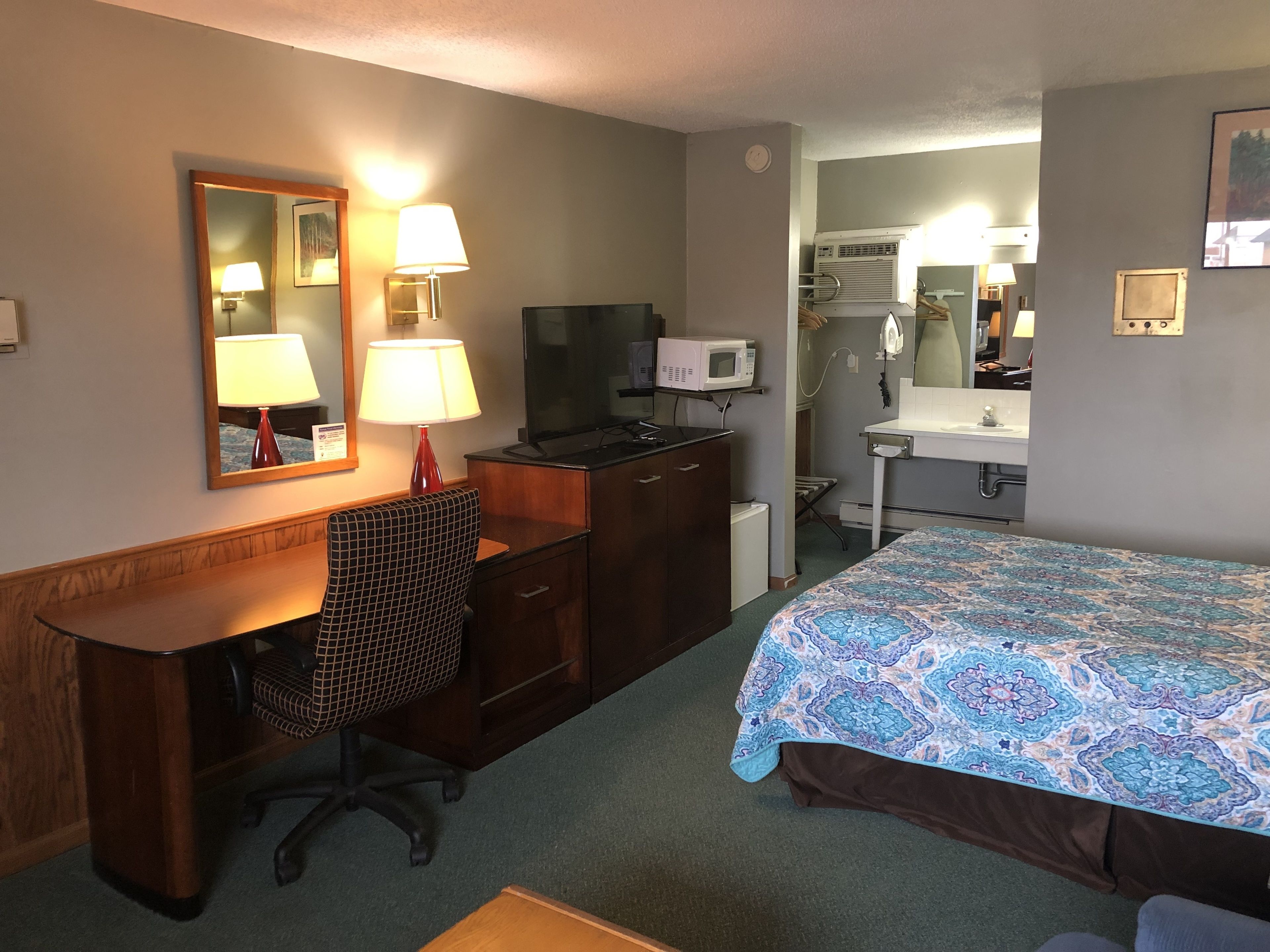 A hotel room with a desk, chair, bed, and a small kitchenette area.