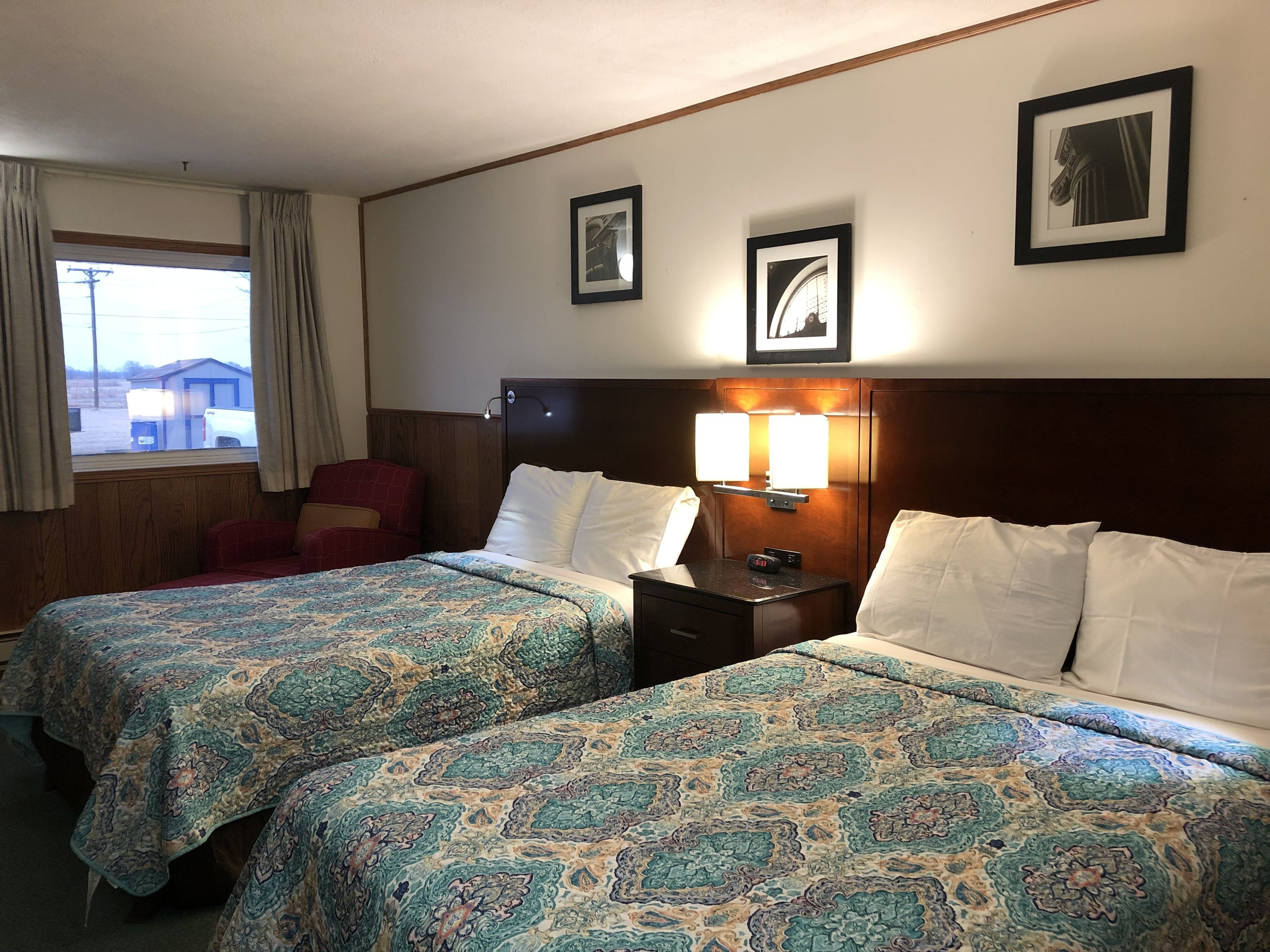 Ein Hotelzimmer mit zwei Betten, einem Nachttisch mit Lampe, einem Sessel und gerahmten Bildern an der Wand.