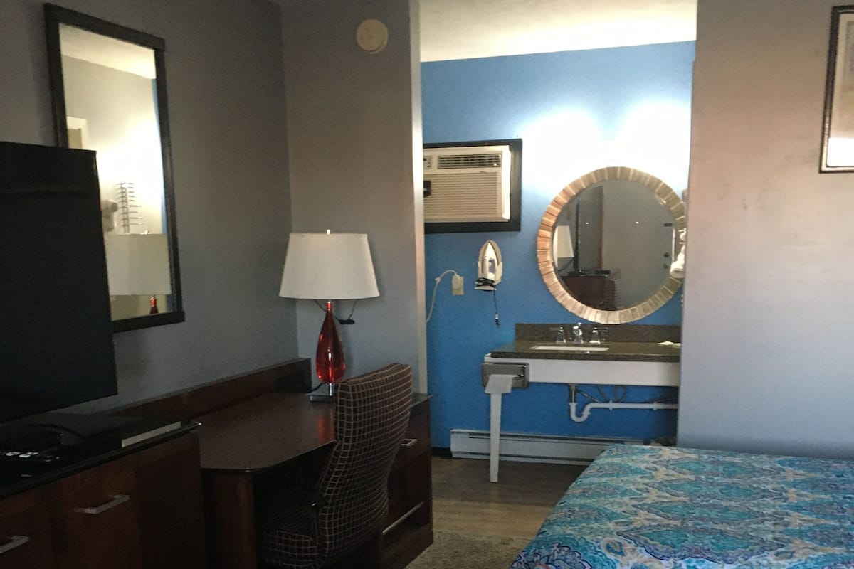 A hotel room with a desk, chair, television, two beds with patterned bedding, a sink with a mirror, and an air conditioning unit.