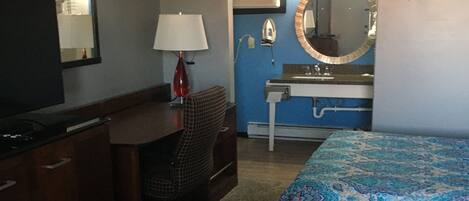 A hotel room with a desk, chair, television, two beds with patterned bedding, a sink with a mirror, and an air conditioning unit.
