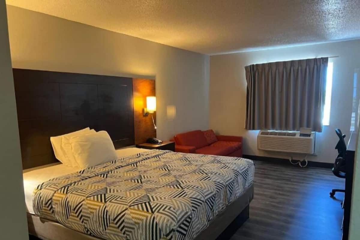 A hotel room with a large bed, a nightstand, a red sofa, a wall-mounted heater, and a window with curtains.