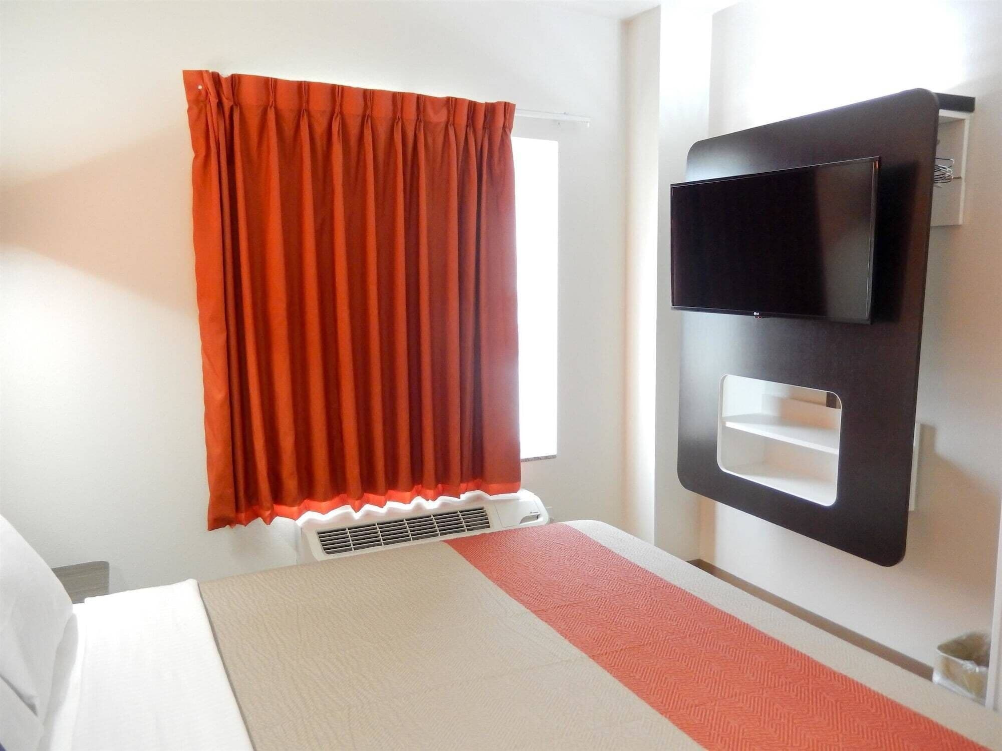 A hotel room with a bed, a TV mounted on the wall, and a window with a red curtain.