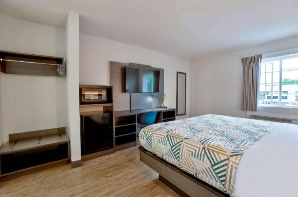 A modern hotel room with a bed, a TV, a desk with a computer, and a window with curtains.