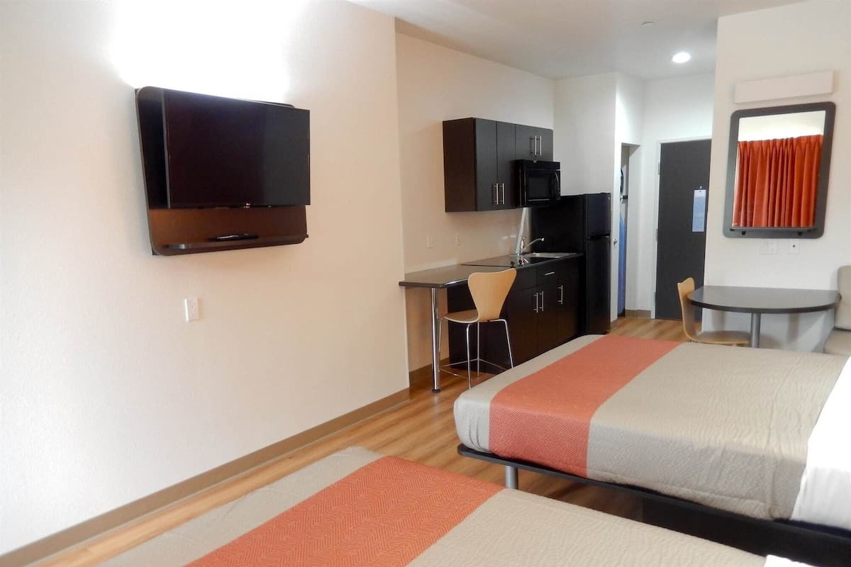 A hotel room with two beds, a desk with a chair, a television, a microwave, a refrigerator, and a small table with a chair.
