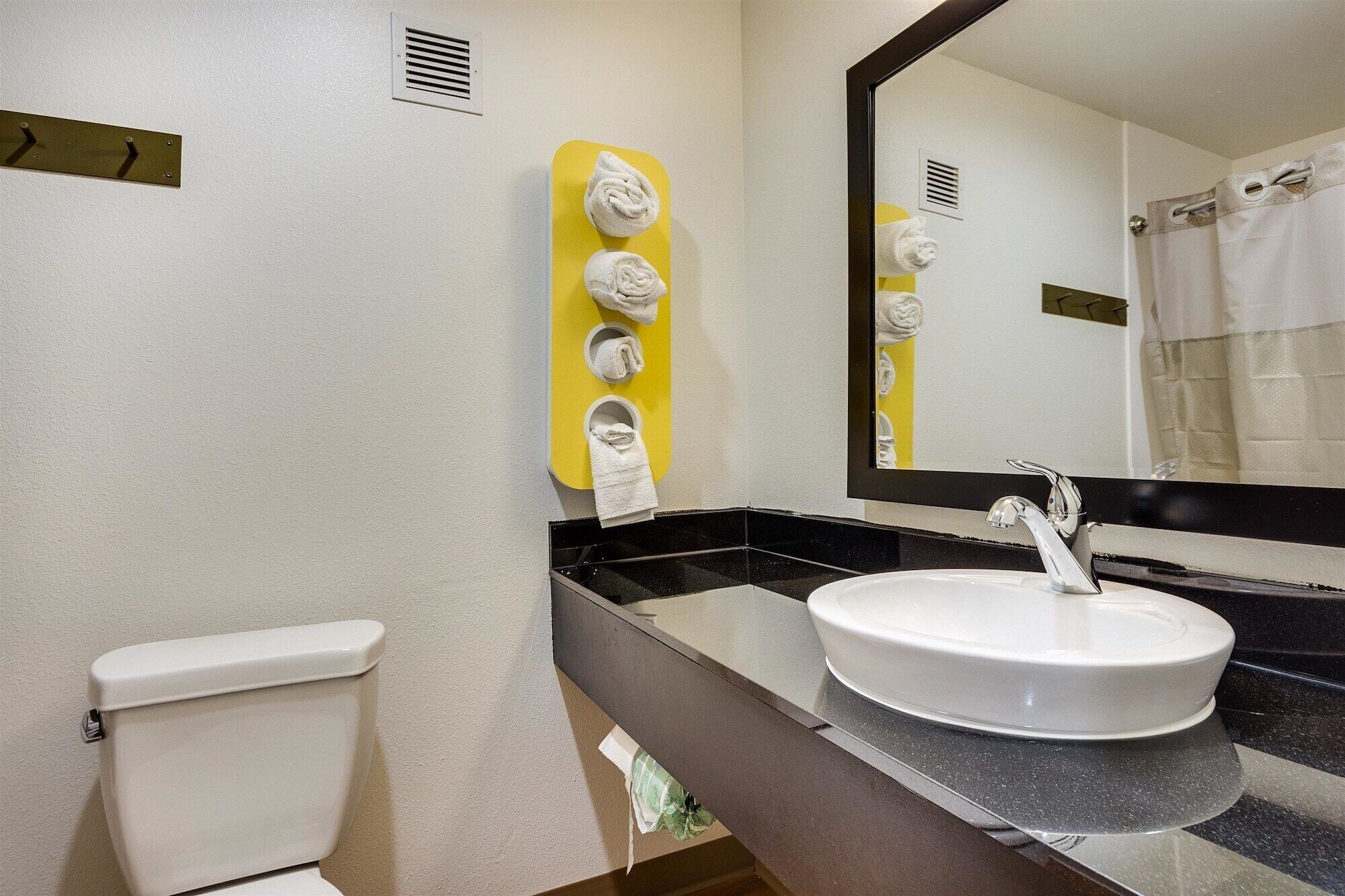 Une salle de bain avec des toilettes blanches, un lavabo avec un robinet chromé, un miroir et un porte-serviettes jaune avec des serviettes.