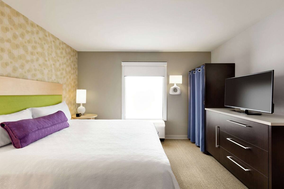 A hotel room with a bed, a television, a nightstand with a lamp, a window with curtains, and a wall-mounted TV.