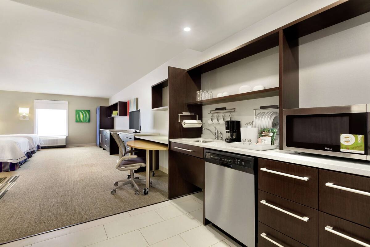 A modern hotel room with a kitchenette, a bed, a desk, and a chair.