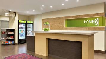 A hotel reception area with a reception desk, a 'HOME 2' sign, a refrigerator stocked with beverages, and a self-service kiosk.