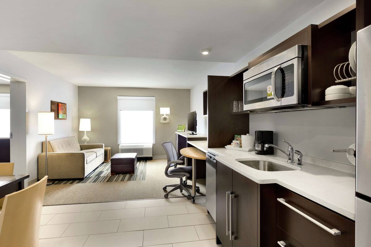 A modern hotel room with a kitchenette, a sofa, a dining table, and a TV.