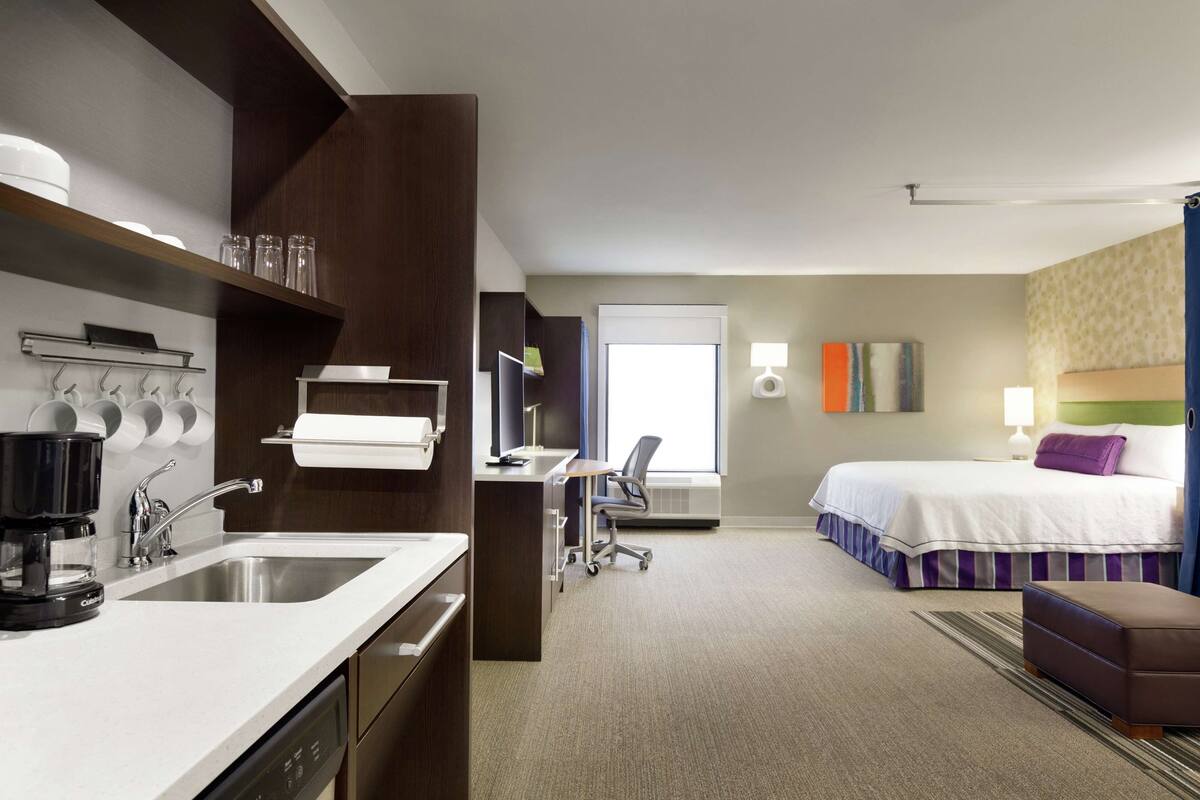 A hotel room with a kitchenette, a bed, a desk, and a chair.