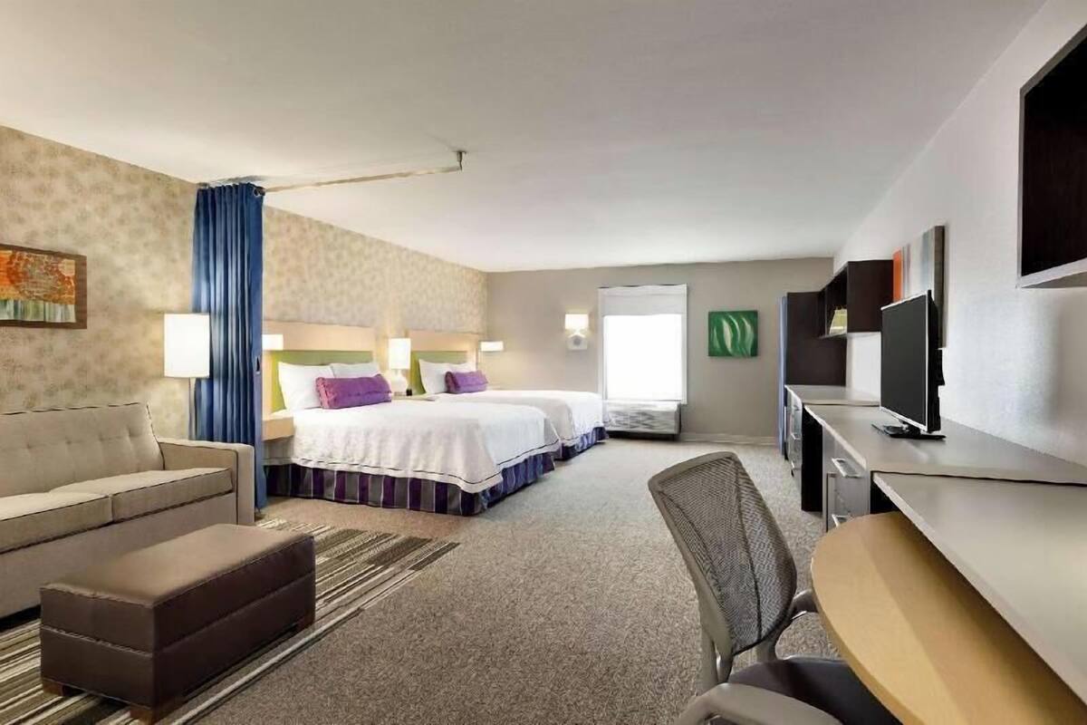 A hotel room with a sofa, two beds, a desk, a TV, and a window.